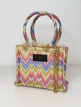 Handgefertigte Mini‑Tote im Zick Zack Muster in der Farbe Ivory/Beige/BLau/Lila/Grün/Gelb/Rosa aus gewebtem Stoff mit seidiger Haptik, etwa 20×17 cm. Sie hat kurze Stoffgriffe, innen rotes Samtfutter, einer abnehmbaren Schulterkette, Magnetverschluss und dekorative Fransenkante.