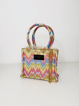 Handgefertigte Mini‑Tote im Zick Zack Muster in der Farbe Ivory/Beige/BLau/Lila/Grün/Gelb/Rosa aus gewebtem Stoff mit seidiger Haptik, etwa 20×17 cm. Sie hat kurze Stoffgriffe, innen rotes Samtfutter, einer abnehmbaren Schulterkette, Magnetverschluss und dekorative Fransenkante.