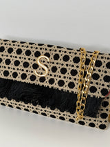 Nahaufnahme der rechteckigen Clutch in schwarz/beige aus handgemachtem, texturiertem Stoff mit edlem gleichmäßigen Punkt Muster und Fransen an der Umschlagklappe. Eine goldfarbene Kette sorgt dafür, dass man die Tasche auch über die Schulter tragen kann. In der Mitte vorne befindet sich das Logo "S" für Soren in Gold. Die Clutch ist vor einem neutralen Hintergrund fotografiert.