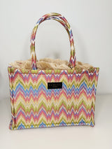 Handgefertigte Medium‑Tote im Zick Zack Muster in der Farbe Ivory/Beige/BLau/Lila/Grün/Gelb/Rosa aus gewebtem Stoff mit seidiger Haptik, etwa 20×17 cm. Sie hat kurze Stoffgriffe, innen rotes Samtfutter, einer abnehmbaren Schulterkette, Magnetverschluss und dekorative Fransenkante.