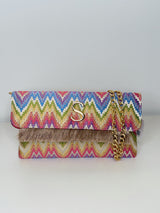 Rechteckige Clutch in abstraktem Zickzack‑Muster in Gelb‑Orange‑Pink-blau-Grün‑Tönen auf naturfarbenem Grund und Fransen an der Umschlagklappe. Eine goldfarbene Kette sorgt dafür, dass man die Tasche auch über die Schulter tragen kann. In der Mitte vorne befindet sich das Logo "S" für Sorena in Gold. Die Clutch ist vor einem neutralen Hintergrund fotografiert.