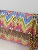 Detailaufnahme der rechteckigen Clutch in abstraktem Zickzack‑Muster in Gelb‑Orange‑Pink-blau-Grün‑Tönen auf naturfarbenem Grund und Fransen an der Umschlagklappe. Eine goldfarbene Kette sorgt dafür, dass man die Tasche auch über die Schulter tragen kann. In der Mitte vorne befindet sich das Logo "S" für Sorena in Gold. Die Clutch ist vor einem neutralen Hintergrund fotografiert.
