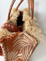 Detailaufnahme der großen rechteckigen Tote‑Bag in Rostrot‑Beige mit gold‑beigem Palmblatt‑Muster aus gewebtem, texturiertem Stoff. Oben Fransenkante, zwei kurze Stoffgriffe, Reißverschluss, rotes Velvet‑Innenfutter, abnehmbare Logo‑Schulterriemen, Maße ca. 42 × 30 × 15 cm