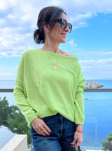 Limettenfarbener Cashmere-Poncho ‚ALTHEA‘ mit weitem U-Boot-Ausschnitt und schmalen Ärmeln, locker fallend, im Hintergrund sieht mas das Meer.
